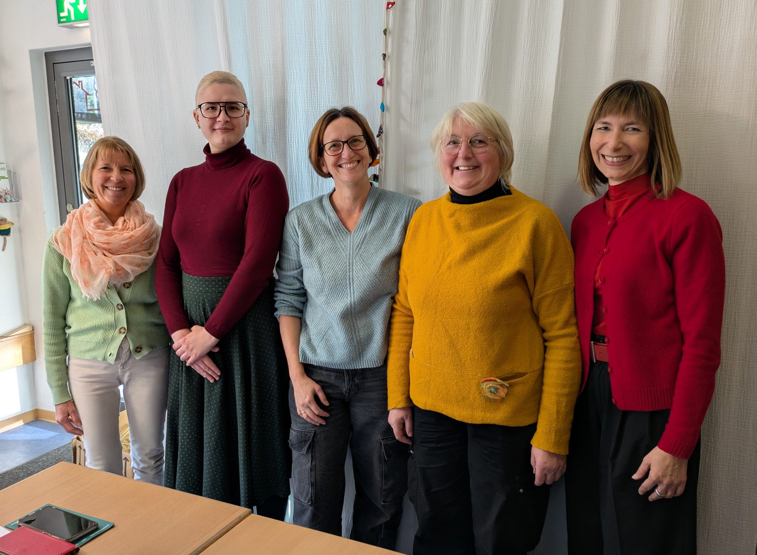 Fünf Frauen lächeln freundlich in die Kamera. Sie tragen farbenfrohe Kleidung uns stehen vor einem weißen Vorhang. Es sind die Deutschlehrerinnen aus den Integrationskursen.