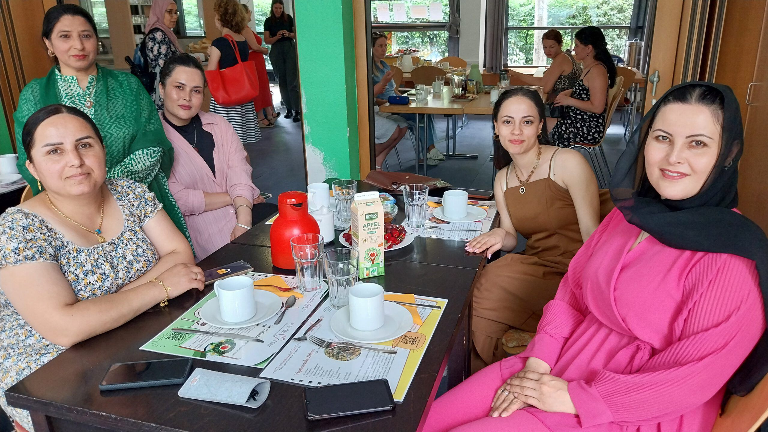 Vier Frauen sitzen an einem Tisch, eine weitere Frau steht daneben. Die Frauen lächeln in die Kamera. Im Hintergrund sind weitere Personen zu sehen.