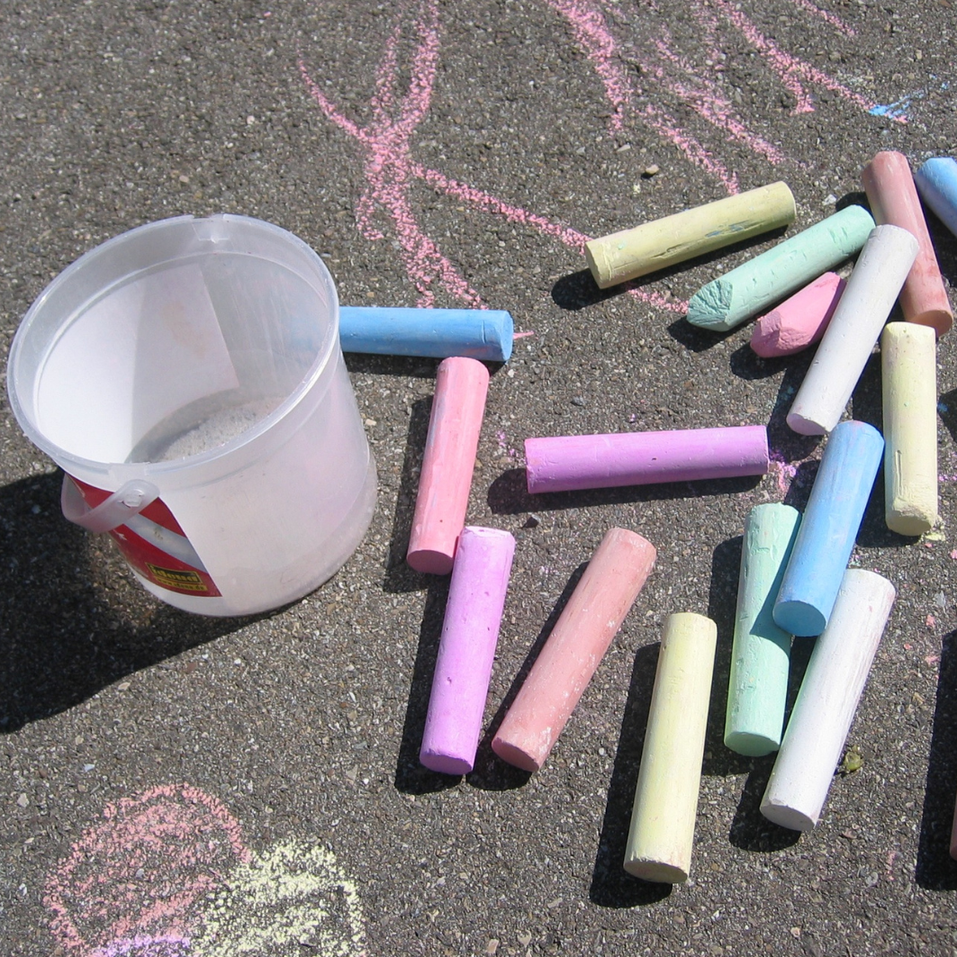 Bunte Straßenkreide liegt auf grauem Asphalt. Links daneben steht ein leerer Plastikeimer.  Der Link führt zu Angeboten für Schulkinder.
