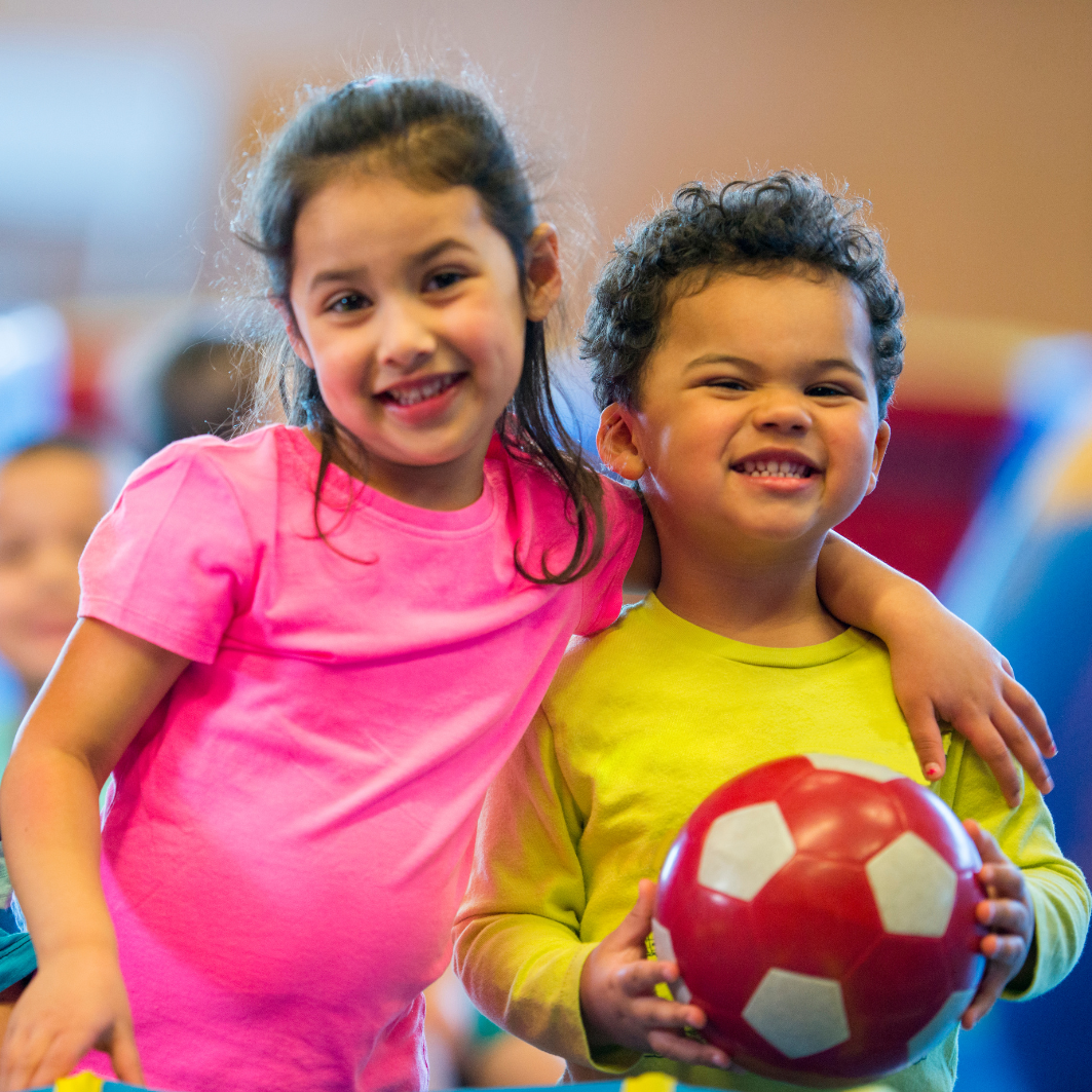Zwei fröhliche Kinder stehen nebeneinander. Ein Mädchen lächelt in die Kamera, sie legt den Arm um einen Jungen neben ihr. Er hält einen roten Ball in den Händen. Der Link führt zu Angeboten für Kinder unter 6 Jahren.