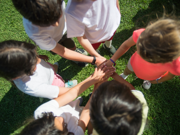 Man sieht von oben eine Gruppe von sechs Kindern, die im Kreis stehen. Sie sind ein Team, jeder legt eine Hand auf die der anderen in der Mitte.