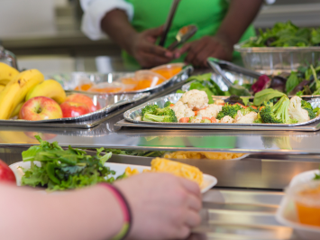 Ein Kind steht an einer Essensausgabe, es gibt frisches Gemüse und Obst.