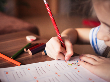 Ein Kind sitzt an einem Tisch und macht Hausaufgaben. In der Hand hält es einen roten Bleistift, Buntstifte liegen neben ihr.
