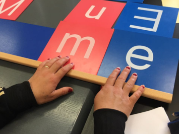 Quadratische Buchstabentafeln aus Holz, links ein rotes M, rechts ein blaues E. Zwei Hände mit lakierten Nägeln liegen auf den Tafeln. Der Link führt zu Informationen zu den Vorkursen.