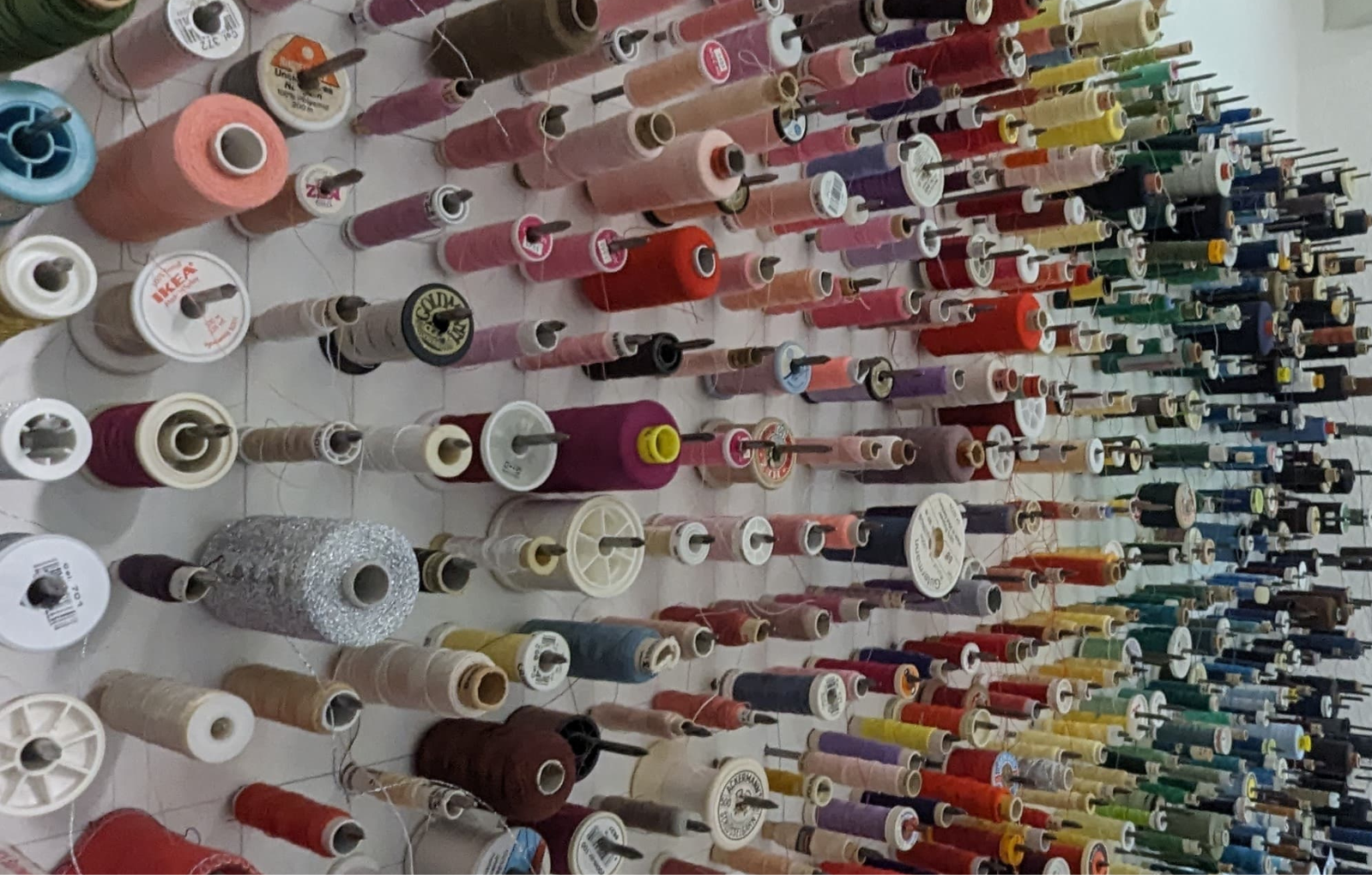 Viele bunte Garnrollen, die an kleinen Stäben an der Wand hängen. Der Link führt auf die Seite des Nähcafés.