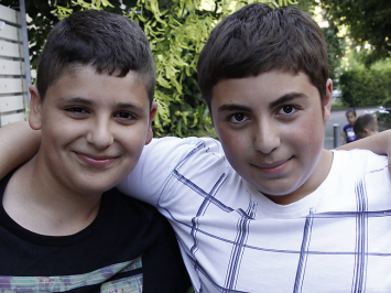 Zwei ungefähr 12 Jahre alten Jungen schauen in die Kamera und lächeln. Sie haben die Arme über die Schultern des anderen gelegt. Der rechte Junge trägt ein weißes Shirt, der linke ein schwarzes.