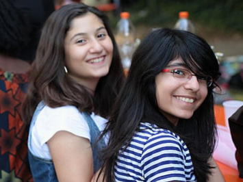 Zwei Mädchen sitzen im Freien. Sie schauen über ihre rechte Schulter lächelnd in die Kamera. Das linke Mädchen trägt ein weißes Shirt und braune Haare. Das rechte Mädchen hat schwarze Haare, eine Brille und einen Pony.