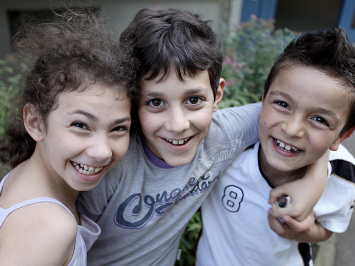 Drei fröhliche Kinder lachen in die Kamera. Sie sind zwischen sieben und zehn Jahren alt. Sie stehen im Freien, hinter ihnen sieht man Blumen.