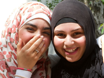 Zwei ungefähr 13 jährige Freundinnen. Das linke Mädchen hält ihre Hand vor das Gesicht, das rechte Mädchen lächelt direkt in die Kamera.