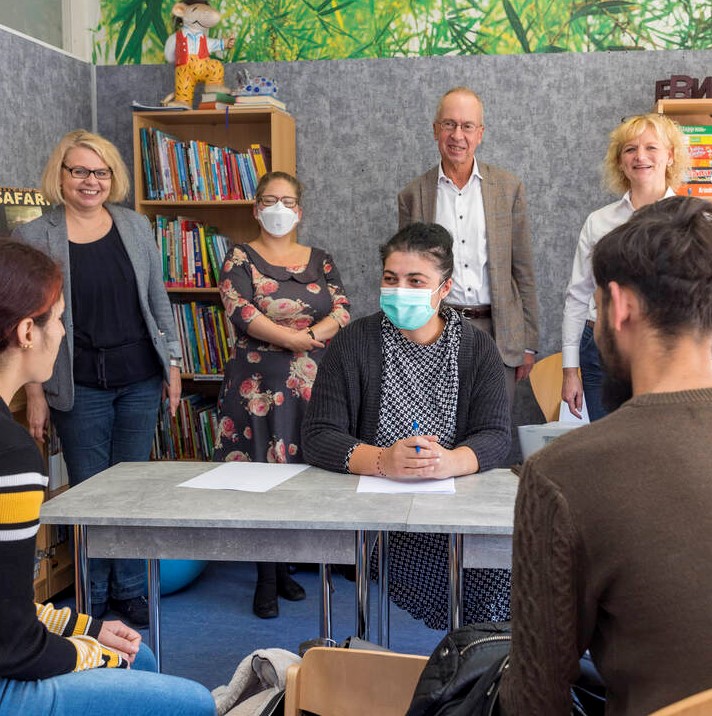 Eine Beratungssituation im Familienzentrum: Eine junge Frau und ein junger Mann sitzen einer Frau gegenüber, die ihnen freundlich entgegenblickt. Zwischen ihnen steht ein Tisch, auf dem Papiere liegen. Hinter der Mitarbeiterin aus der Beratung stehen weitere Personen, die in die Kamera lächeln.