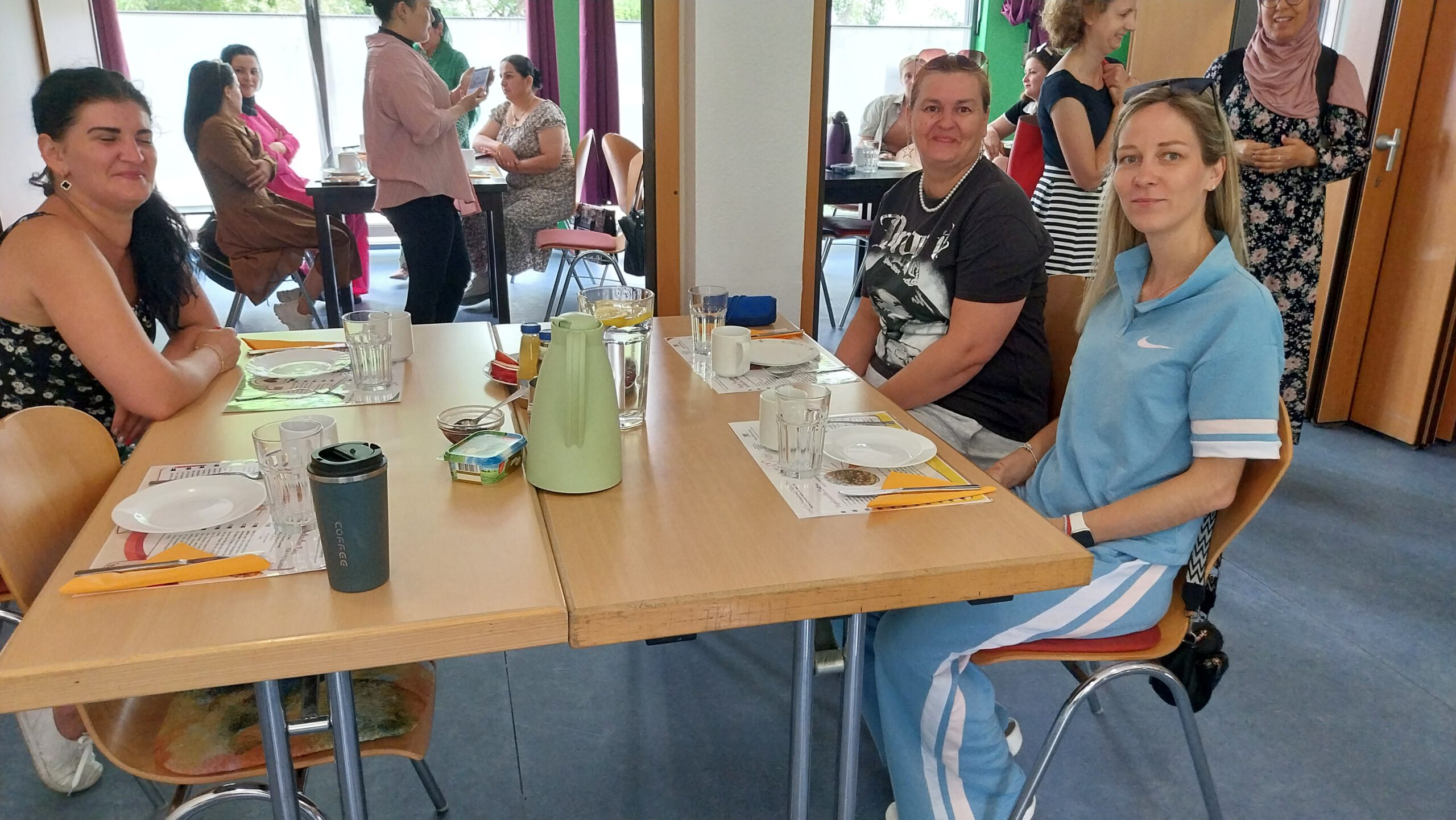 Drei Frauen sitzen an einem Tisch, der Tisch ist für das Essen gedeckt. Im Hintergrund sind weitere Frauen, die sich unterhalten.