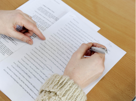 Eine Person bearbeitet einen langen Text, der mit dem Computer geschrieben wurde. Er steht für die Fähigkeit "Schreiben". Der Link führt zum Menüpunkt Deutschkurse.