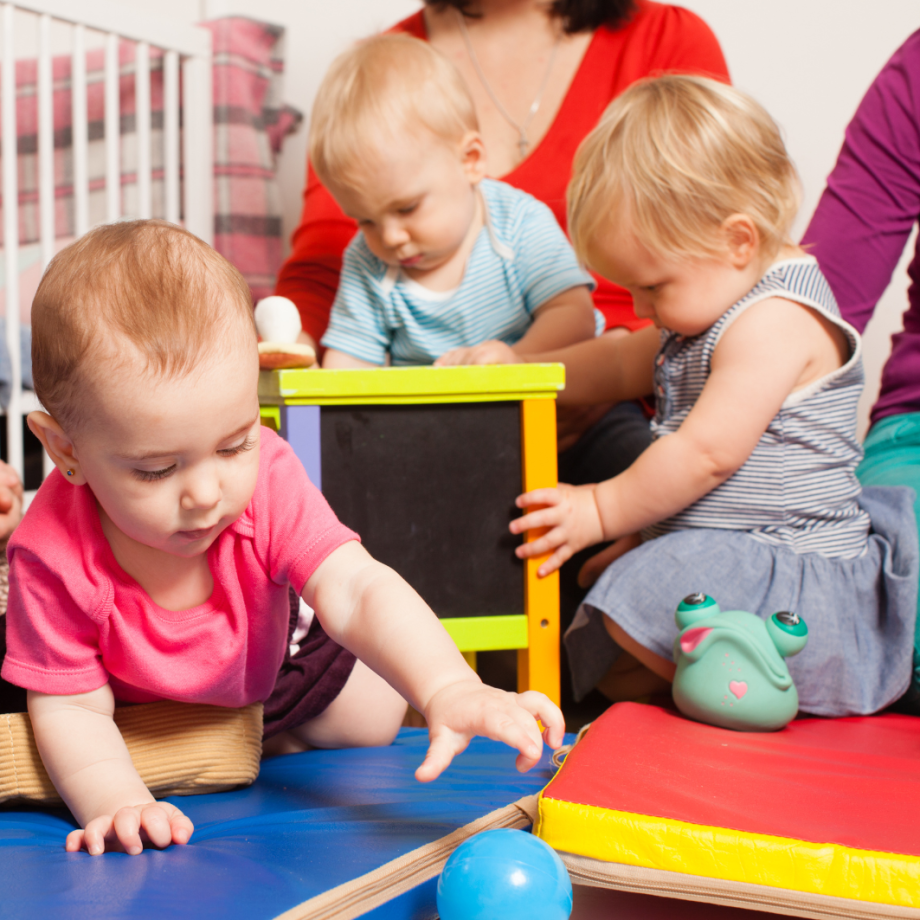 Ein ungefähr einjähriges Baby mit einem pinken Shirt greift nach einem blauen Ball. Im Hintergrund untersuchen zwei weitere Kleinkinder eine Tafel. 