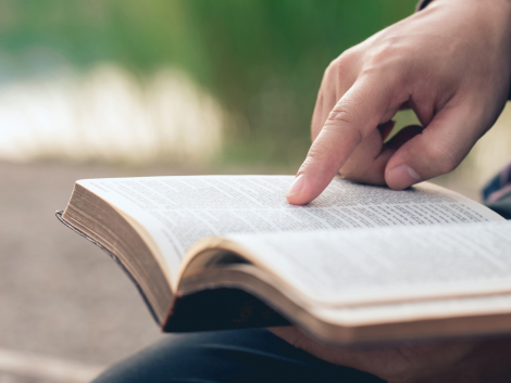 Eine Person hat ein aufgeschlagenes Buch auf den Knien liegen, der Zeigefinger liegt auf dem Buch. Das Bild steht für die Fähigkeit Lesen.