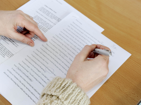 Eine Person bearbeitet einen langen Text, der mit dem Computer geschrieben wurde. Er steht für die Fähigkeit "Schreiben". Der Link führt zum Menüpunkt Deutschkurse.