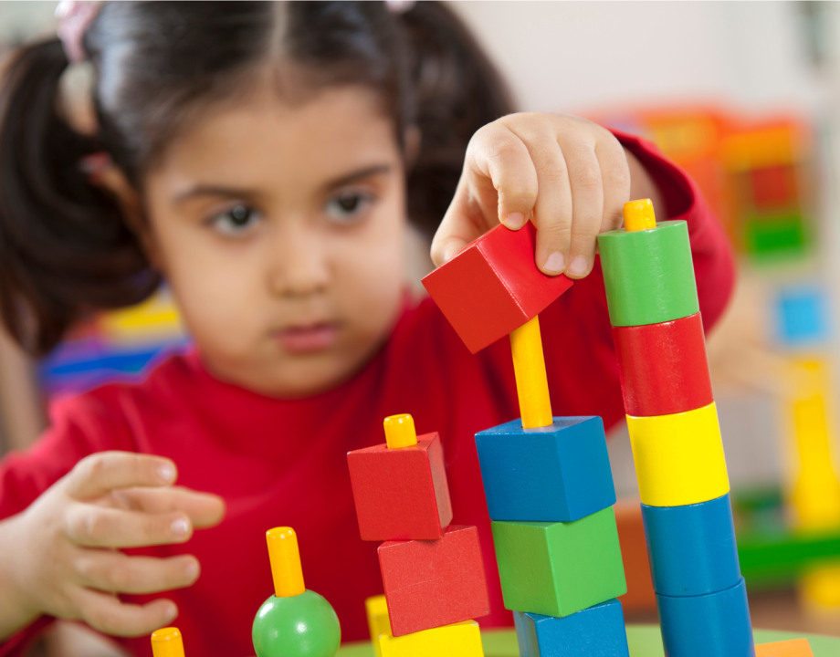 Ein ungefähr dreijähriges Kind macht sehr konzentriert ein Steckspiel mit bunten Holzklötzen. Sie trägt zwei Zöpfe und einen roten Pullover.