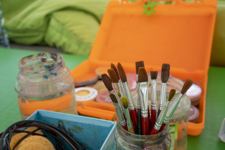 Ein Glas mit vielen Pinseln, im Hintergrund eine orangene Dose mit Bastelmaterial.