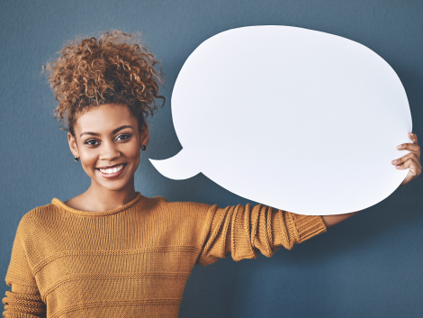 Eine Frau mit lockigen Haaren blickt lachend in die Kamera, sie hält eine leere Sprechblase wie aus einem Comic neben ihrem Kopf. Das Bild steht für die Fähigkeit Sprechen.